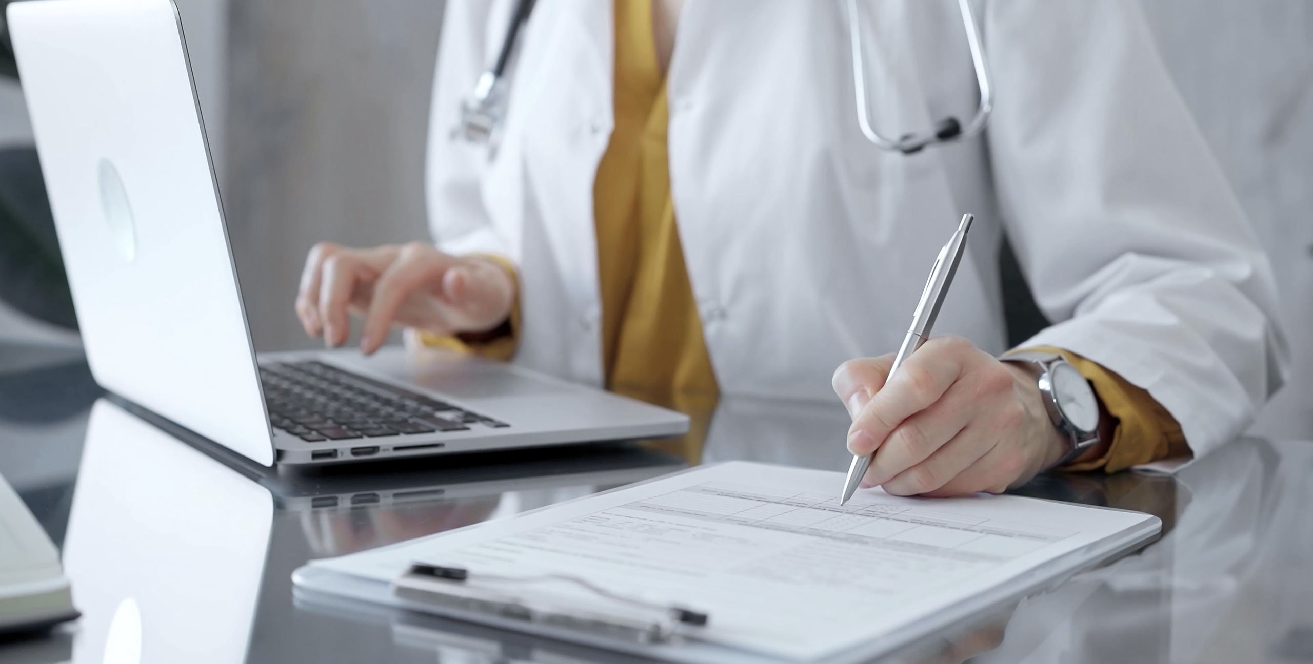 Doctor woman is writing on a document on a glass desk, with a laptop and tablet visible. Work space concept in Medicine